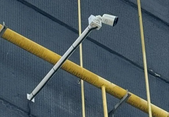 Камера видеонаблюдения на длинном выносном кронштейне BOXFORCAM™ на тёмной кирпичной стене промышленного корпуса; наискось по стене проходит жёлтая газовая труба на настенных держателях, кабель уложен по внешней части кронштейна