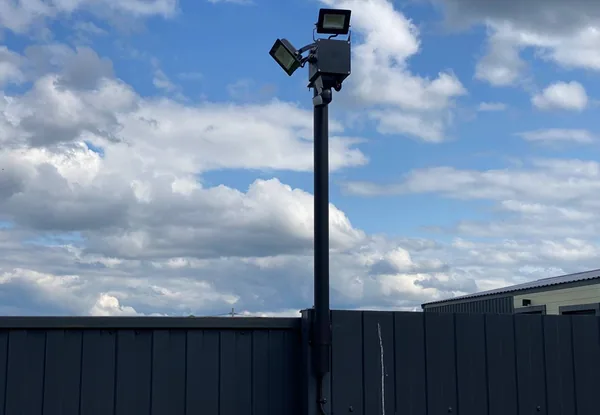 Мачта BOXFORCAM™ поднята над металлическим забором участка, наверху два прожектора и датчик движения, фон — небо